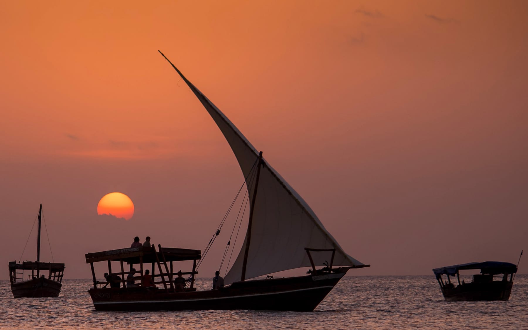 Dhows sailing at sunset with people silhouetted on the shore